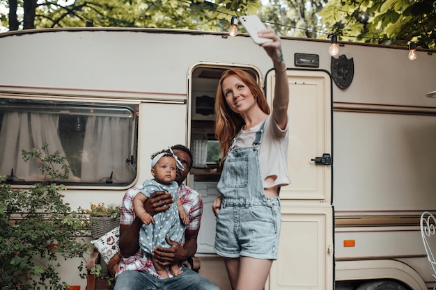 Portrait of candid happy interracial family with swarthy baby take selfie near retro travel trailer