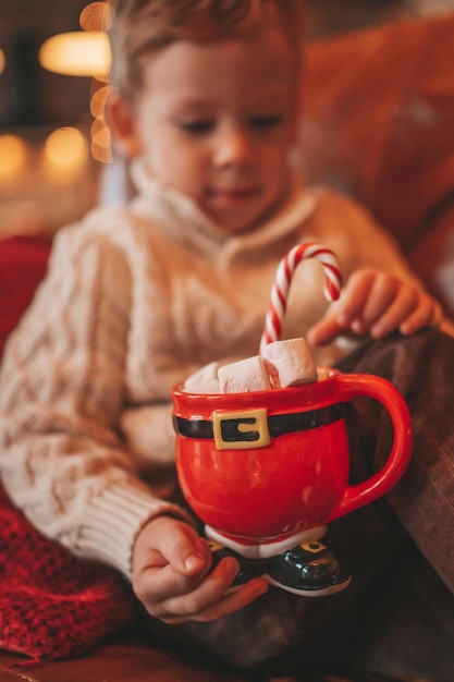 Portrait candid happy kid in knit beige sweater hold Xmas mug with marshmallows and candy cane