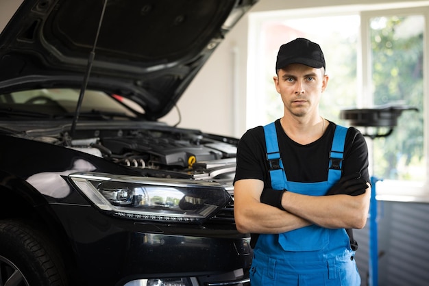 Portrait of a car mechanic crosses hands in a car workshop in blue uniform with equipment looking