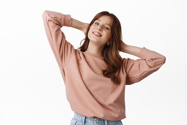 Portrait of carefree redhead woman relaxing on weekend, holding hands behind head and smiling, resting and having fun, standing over white background