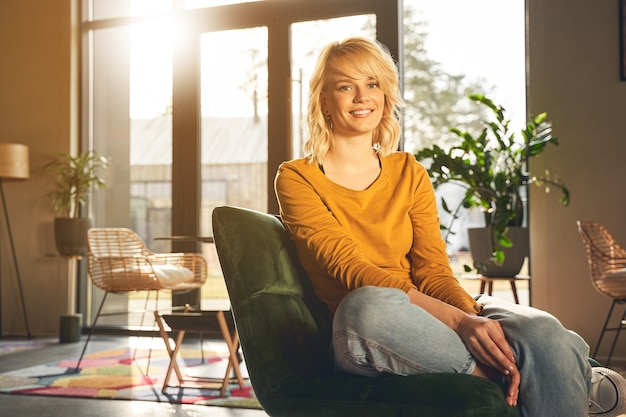 Portrait of a carefree young blonde Caucasian lady in casual clothes sitting in an armchair