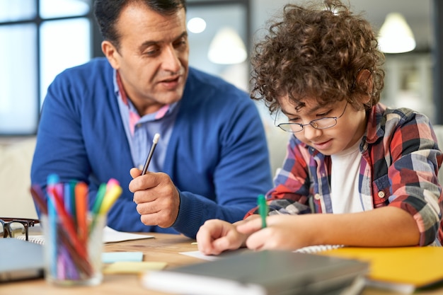 Portrait of caring middle aged latin father helping, discussing homework with his son, school child while sitting together at the desk at home. Remote learning, family, fatherhood concept