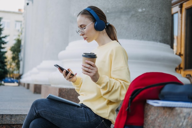 Portrait of casual student girl in headphones using mobile phone app outdoors young woman student