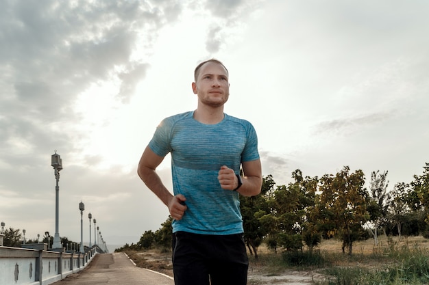 Portrait of Caucasian guy in a blue t-shirt and black shorts who trains and runs on the asphalt track during sunset