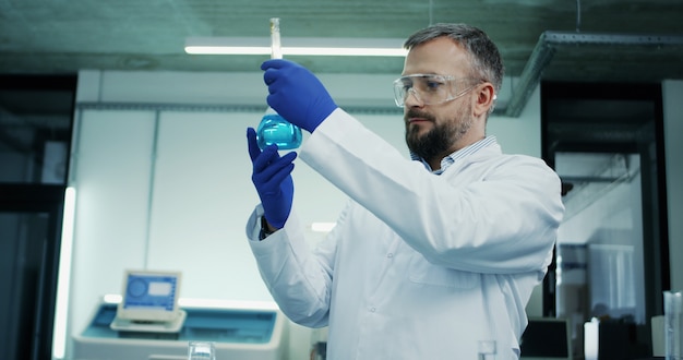 Portrait of the Caucasian man in glasses and white robe doing an analysis of the blue liquid in the test tube during medical or pharmaceutical research in the laboratory.