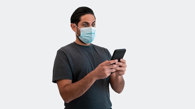 Portrait of caucasian man raising eyebrows because of surprise with smartphone and medical mask during corona virus outbreak. Surprised reaction person with phone device on white background