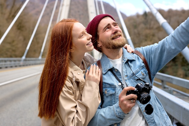 Portrait of caucasian man and woman in casual wear walking on bridge, in contemplation of mountains, landscapes around. Handsome male pointing finger at side, showing something to girlfriend