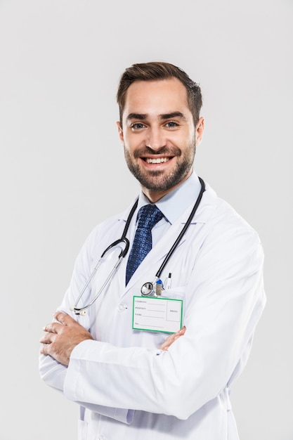 Portrait of caucasian young medical doctor with stethoscope smiling and standing wth arms crossed isolated over white wall