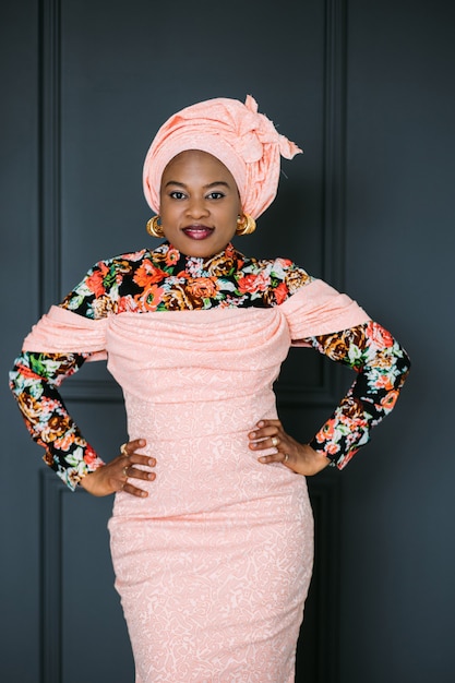 Portrait of charming African woman wearing stylish pink dress and turban on head, smiling on camera with arms on waist, posing on dark blue wall background.