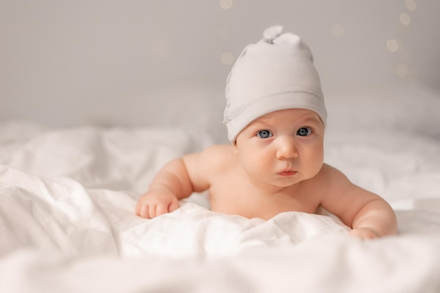 Portrait of a charming cheerful baby with blue eyes in a white cap lying on his tummy on white bed