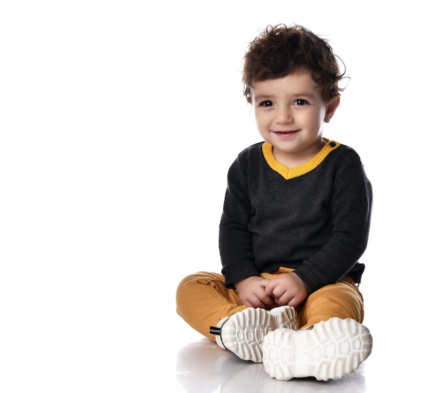 Portrait of the charming curly baby with big brown eyes which sits on a white background and looking up smiling The concept of stylish clothes for children