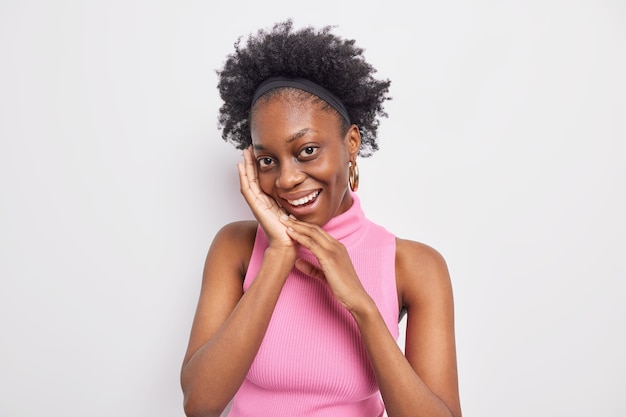 Portrait of charming dark skinned woman with natural beauty keeps hands near face looks gently at camera