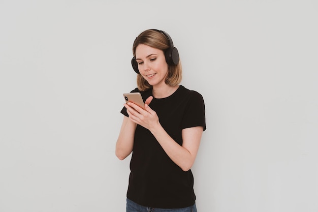 Portrait of charming European woman in casual clothes with headphones listening to music on smartphone Isolated on white background.