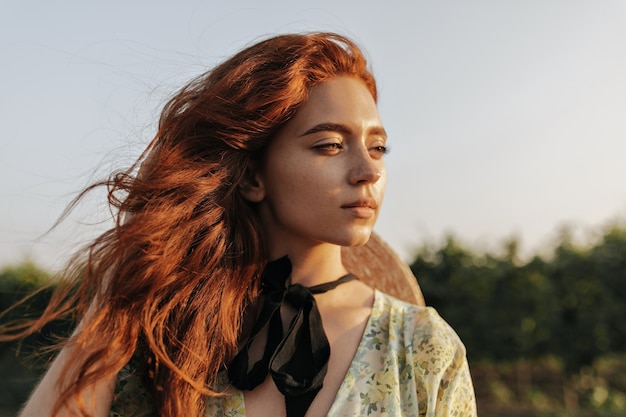 Portrait of charming girl with bright ginger hair, lovely freckles and dark bandage on neck looking away and posing outdoor