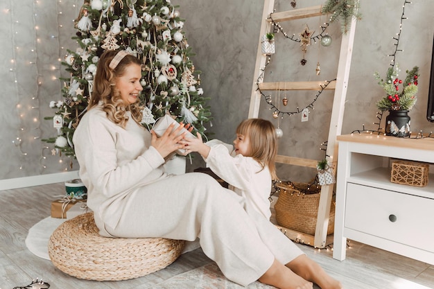 Portrait of a charming little girl receiving a gift from a loving mother, sitting near the Christmas tree. Beautiful woman makes a New Year's gift to her child.