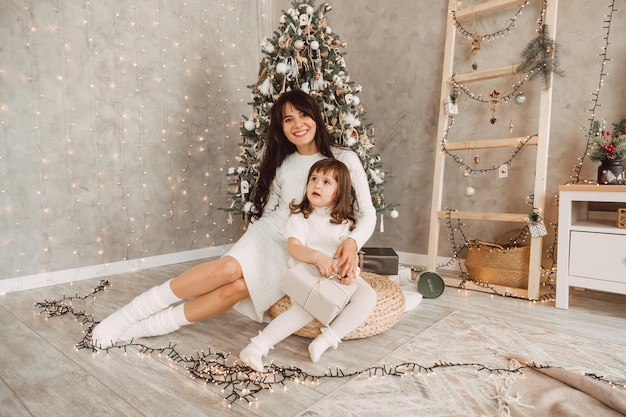 Portrait of a charming little girl receiving a gift from a loving mother, sitting near the Christmas tree. Beautiful woman makes a New Year's gift to her child.