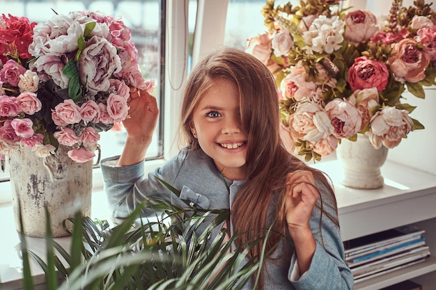 Portrait of a charming little girl with long brown hair and piercing glance wearing a stylish dress, smiling while posing with flowers against a window at home