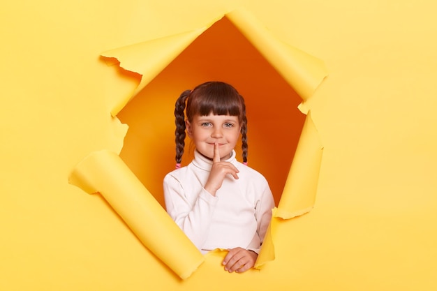 Portrait of charming smiling little girl with braids wearing white turtleneck keeps finger near lips asking to not telling secret posing in torn hole of yellow paper wall