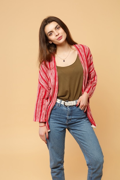 Portrait of charming young woman in casual clothes looking camera, holding hand in pockets isolated on pastel beige background in studio. People sincere emotions lifestyle concept. Mock up copy space.