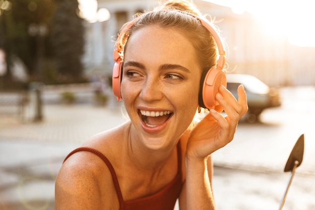 Portrait of charming young woman listening to music with wireless headphones while sitting on scooter outdoors on city street