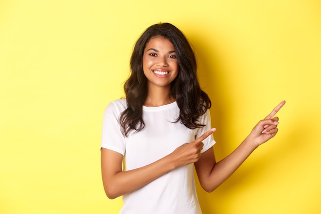 Portrait of cheerful african-american teenage girl, smiling happy and pointing fingers right at information banner, showing copy space, standing over yellow background.