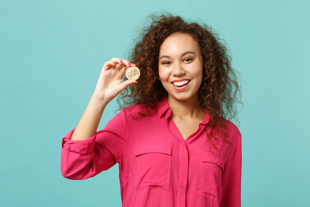 Portrait of cheerful african girl in casual clothes holding bitcoin future currency isolated on blue turquoise wall background in studio. People sincere emotions lifestyle concept. Mock up copy space.