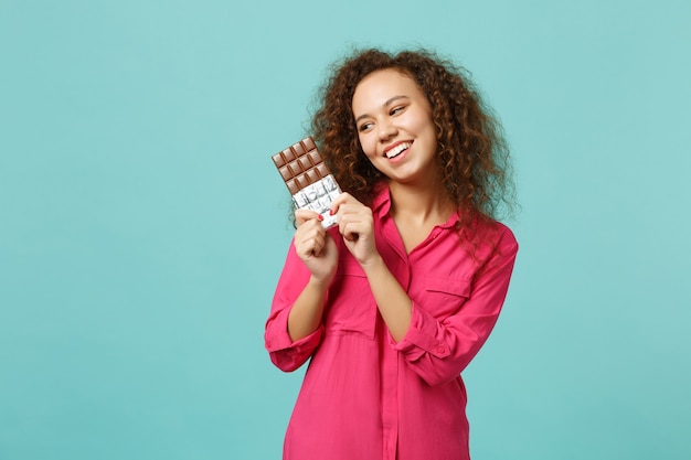 Photo portrait of cheerful african girl in casual clothes holding in hand chocolate bar isolated on blue turquoise wall background in studio. people sincere emotions, lifestyle concept. mock up copy space.