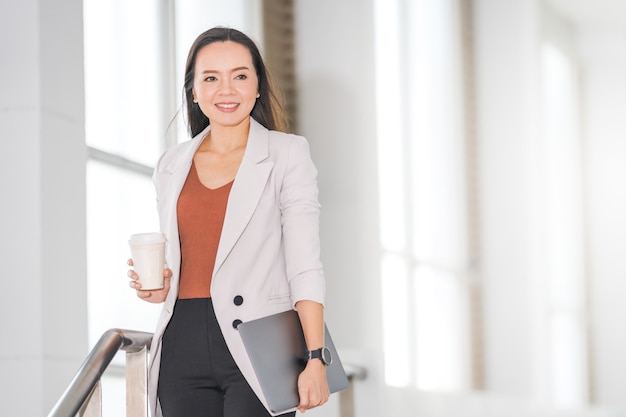 Portrait Of Cheerful Asian Business Woman Holds Take Away Coffee And Digital Tablet in Company