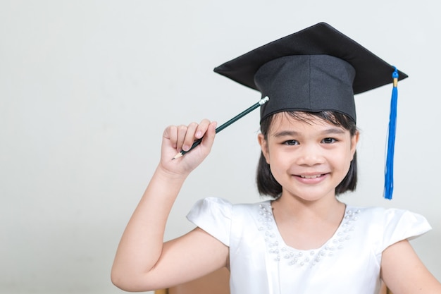 Portrait of cheerful Asian little girl student wear a mortarboard or graduation hat hold pencil and write on notebook with white background with copy space. Education Graduation Concept Stock Photo