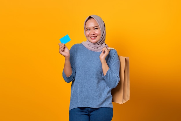 Portrait of cheerful Asian woman holding shopping bag and showing credit card over yellow background