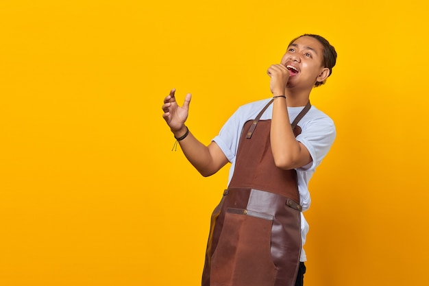 Portrait of cheerful asian young wearing an apron is seen enjoying singing on yellow background