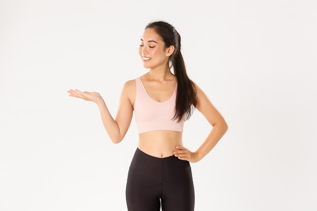 Portrait of cheerful attractive asian fitness coach, female athlete introduce or demonstrate product for workout, holding something on hand.