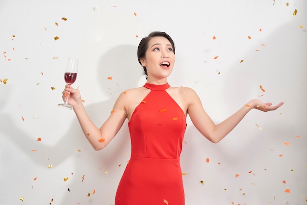 Portrait of a cheerful beautiful girl wearing dress standing under confetti rain and celebrating isolated over white background