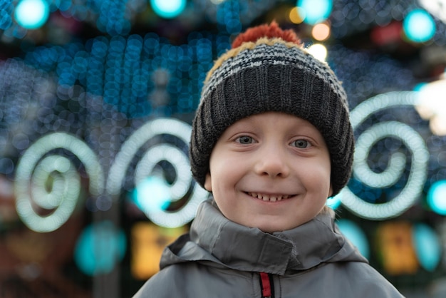 Portrait of cheerful child boy in knitted hat on street Christmas decorations on the background