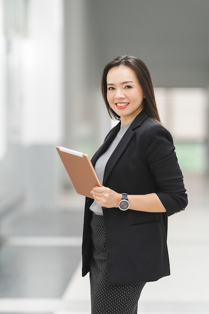 Portrait of a cheerful confident Asian businesswoman in a business suit standing whie using a digital tablet in the business building. Business stock photo