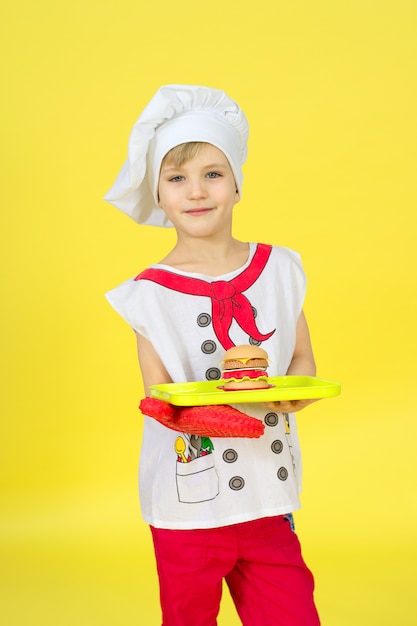 Portrait of a cheerful cook boy with a burger on a tray on a yellow wall. Chef in red uniform. Different occupations