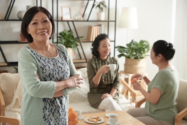 Portrait of cheerful elegant senior woman standing in living room her friends drinking tea and chatt...