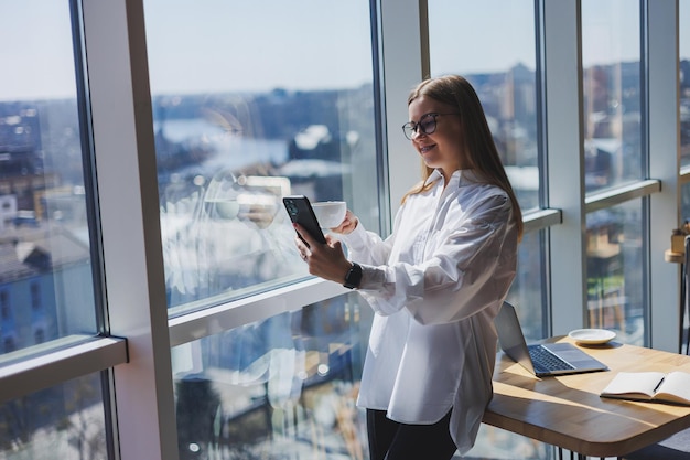 Portrait of a cheerful european blogger woman in glasses smiling while chatting online using her phone joyful woman in a white shirt and a cup of coffee posing in a cafe