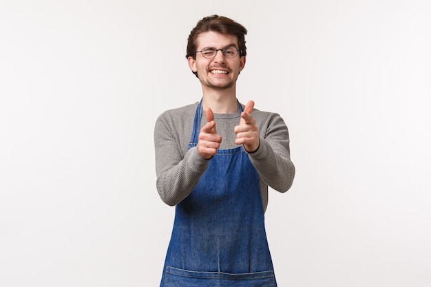 Portrait of cheerful friendly man in apron, work at coffee shop or restaurant, show informal greeting sign finger pistols pointing and wink, inviting to visit his store, white wall