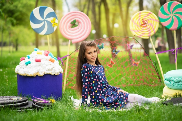 Portrait of cheerful girl on the green grass in the scenery from huge sweets of lollipops and cakes