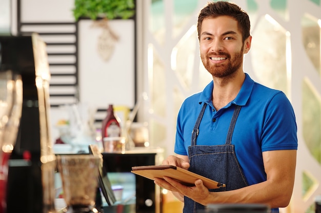 Portrait of cheerful handsome coffeeshop waiter standing with tablet computer in hands and smiling at camera