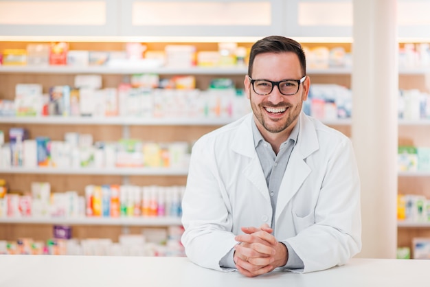 Portrait of a cheerful handsome pharmacist leaning on counter at drugstore.