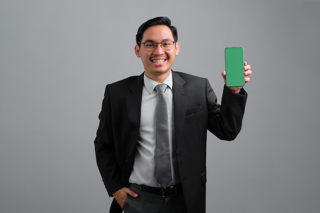 Portrait of cheerful handsome young businessman in formal suit showing smartphone with empty screen isolated on grey background