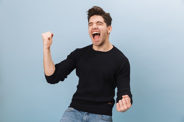 Portrait of a cheerful handsome young man standing isolated on blue, celebrating success