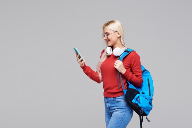 Portrait of a cheerful happy blond female student with blue backpack listening online lecture on phone with headphones isolated over grey space. Copy space for text on the left side