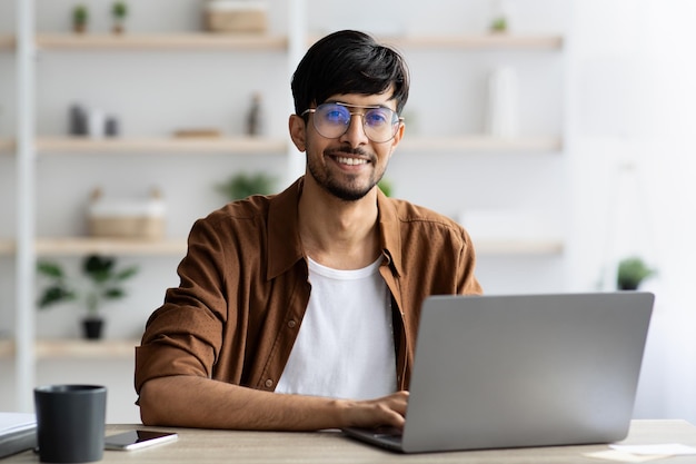 Portrait of cheerful indian guy working on laptop