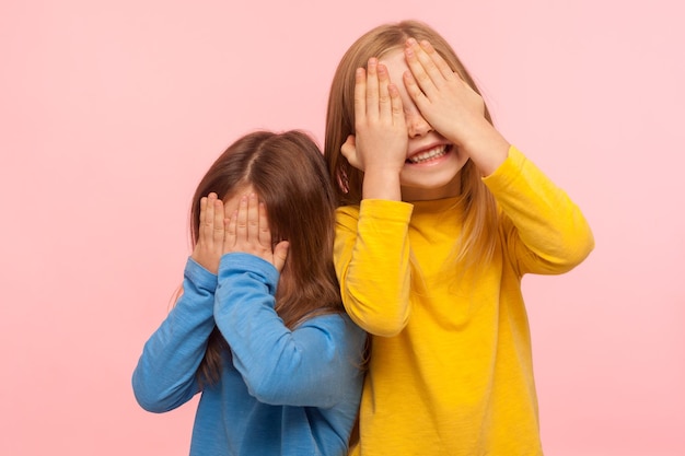 Photo portrait of cheerful joyful carefree little girls covering their faces with arms and sincerely smiling, laughing, having fun and playing peek a boo game. indoor studio shot isolated on pink background