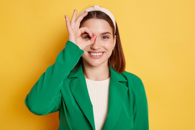 Portrait of cheerful joyful woman in green jacket and hair band posing isolated over yellow background looking at camera with toothy smile and covering eye with ok sign showing approving gesture