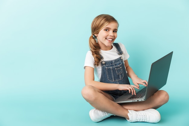 Portrait of a cheerful little girl isolated over blue wall, studying with laptop computer