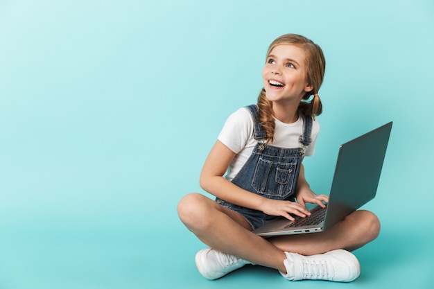 Portrait of a cheerful little girl isolated over blue wall, studying with laptop computer